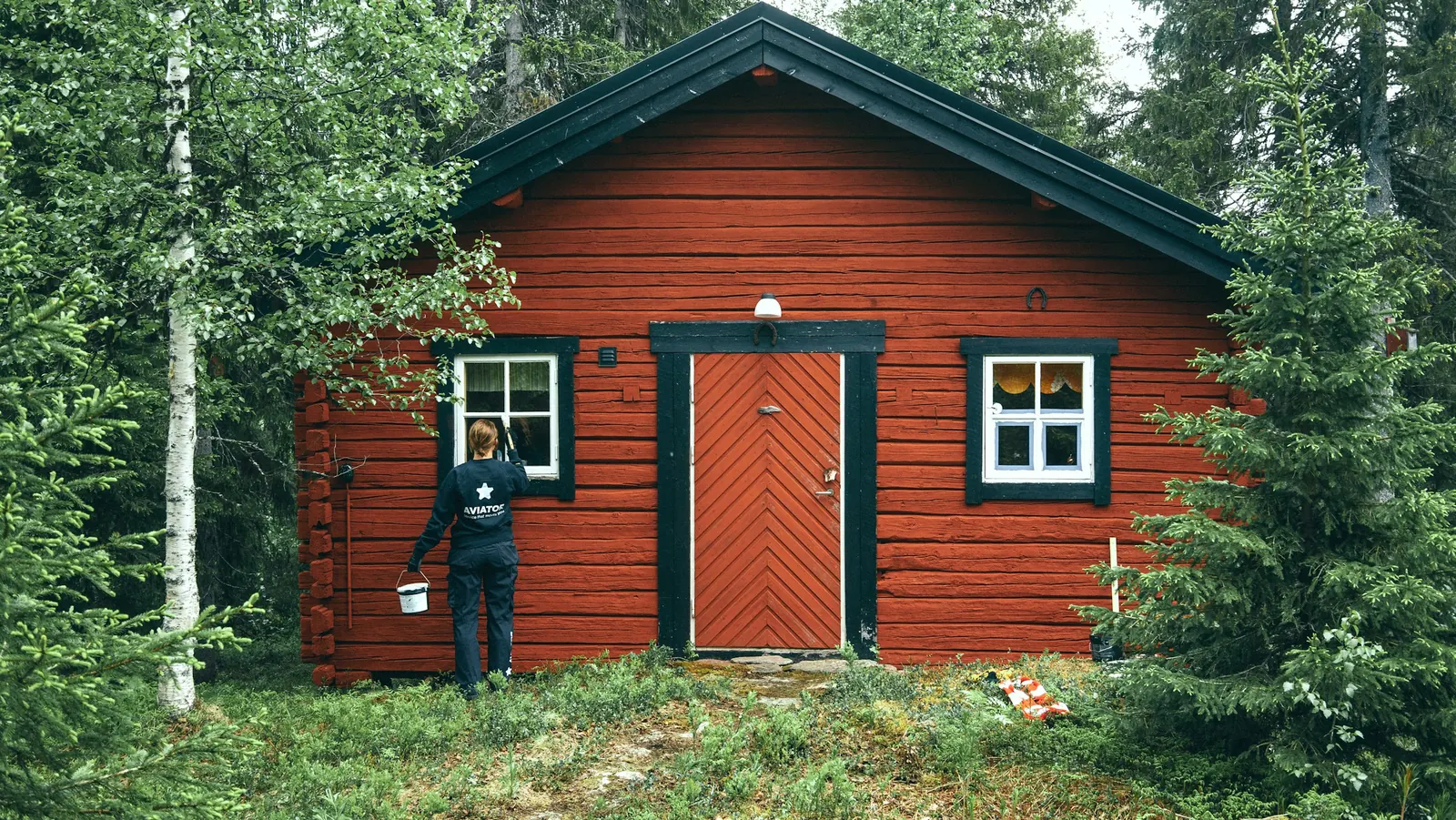 Målare framför en nymålad röd timmerstuga i skogen